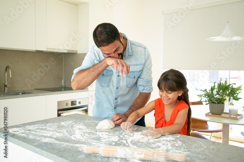 Happy dad and daughter having fun while kneading dough on kitchen table. Father teaching his girl to bake bread or pies. Family cooking concept