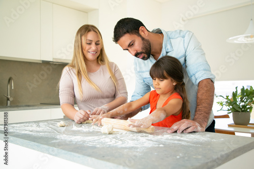 Positive parents watching daughter rolling dough on kitchen desk with flour messy. Young couple and their girl baking buns or pies together. Family cooking concept