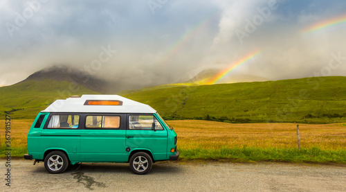 Fototapeta Naklejka Na Ścianę i Meble -  An old green camper van in the shadow of misty mountains and a rainbow