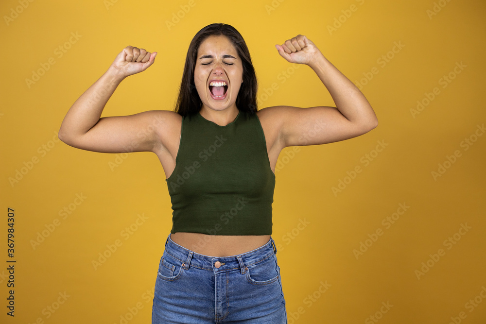 Young beautiful woman standing over yellow isolated background screaming with her arms in the air with a gesture of victory Young beautiful woman standing over yellow isolated background screaming with her arms in the air with a gesture of victory
