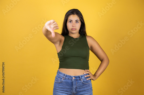 Young beautiful woman standing over yellow isolated background giving thumb down gesture looking with negative expression and disapproval