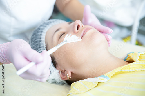 woman is lying in the cosmetologist s office on the procedure of moisturizing her face. The cosmetologist applies a moisturizing mask to the patient's face.