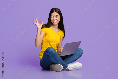 Everything ok. Joyful asian girl sitting with laptop and showing okay sign
