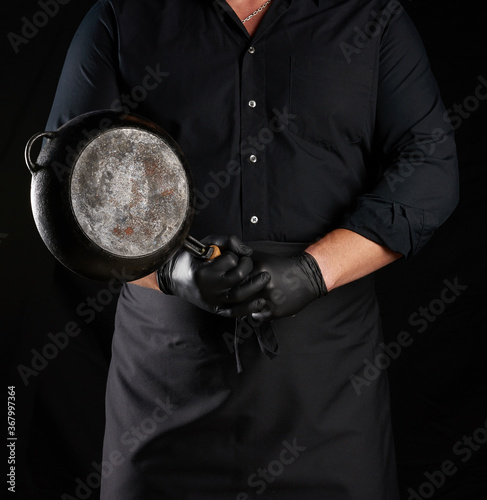male cook in black uniform and latex gloves holds an empty round vintage black cast iron pan in front of him