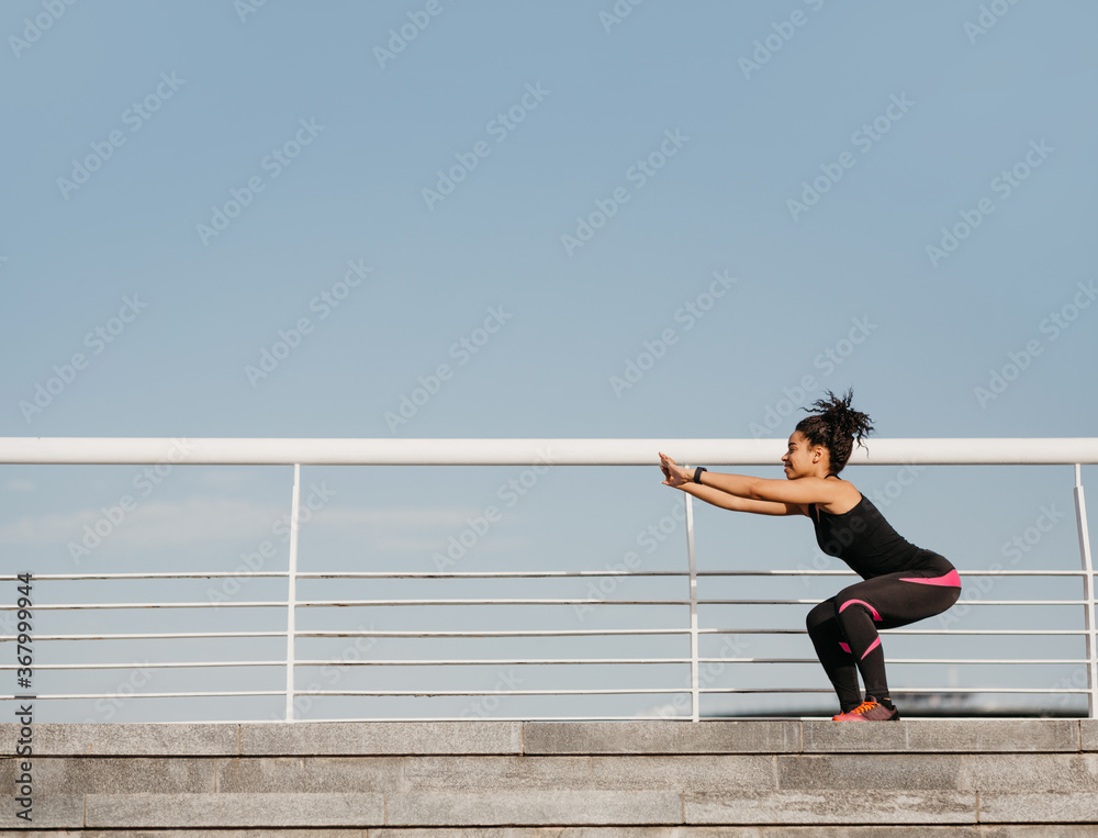 Preparing for marathon outdoors. African american girl in sportswear with fitness tracker doing squats Preparing for marathon outdoors. African american girl in sportswear with fitness tracker doing squats