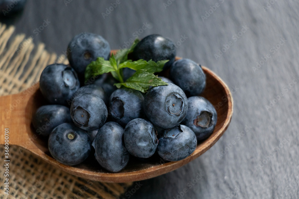 Several blueberries and green mint leaves on a wooden spoon and a burlap napkin. Black stone background. Healthy snack. Several blueberries and green mint leaves on a wooden spoon and a burlap napkin. Black stone background. Healthy snack.