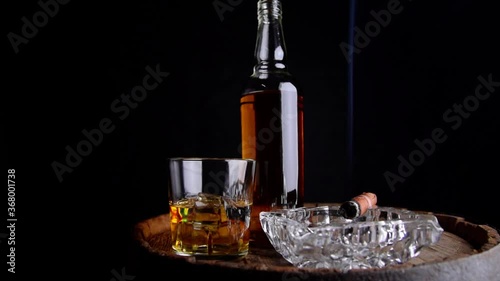A glass of whiskey with ice cubes, a bottle and a cigar in the smoke on a wooden barrel