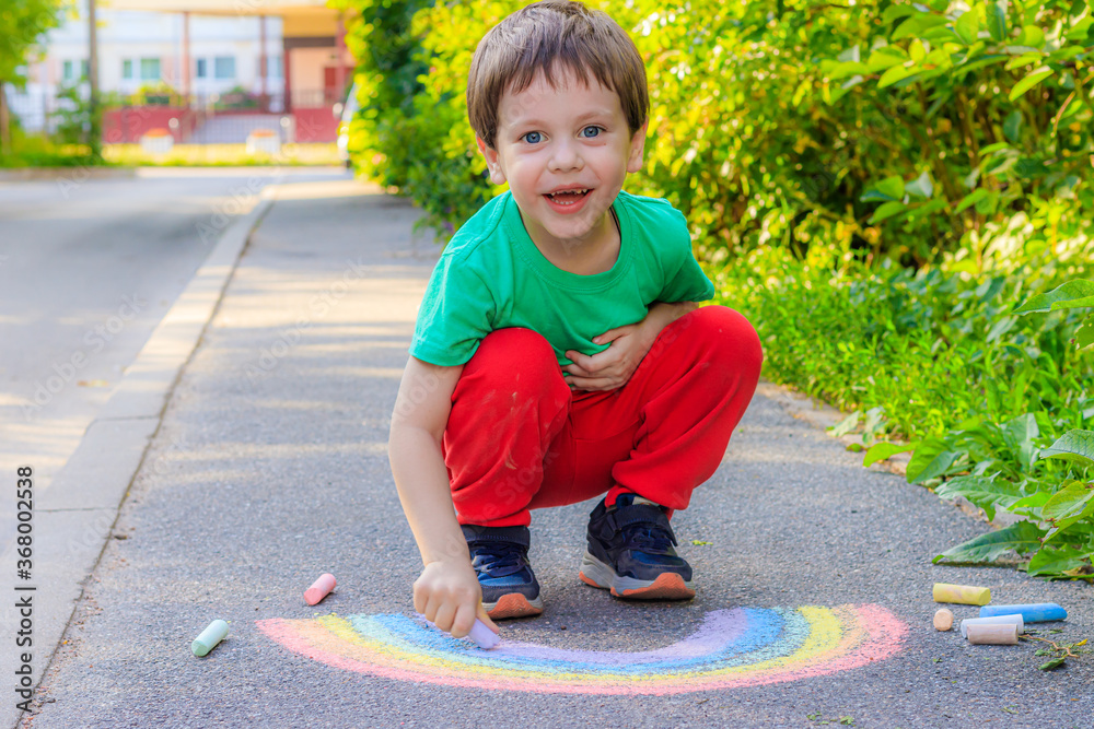 Fototapeta premium A boy draws a rainbow on the asphalt with crayons . A child draws on the asphalt . Children's drawing. Walking in the city with a child. Summer in the city. Crayons for drawing.