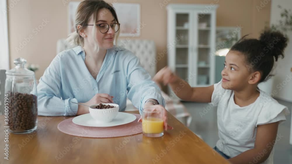 Breakfast of multiethnic family in modern cozy kitchen. At home honey African preschool daughter thoughtfully eats corn flakes drinking orange juice. Girl plays with food making caring mother laugh. Breakfast of multiethnic family in modern cozy kitchen. At home honey African preschool daughter thoughtfully eats corn flakes drinking orange juice. Girl plays with food making caring mother laugh.