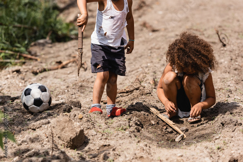 Selective focus of destitute african american children playing with wooden twigs near soccer ball on urban street