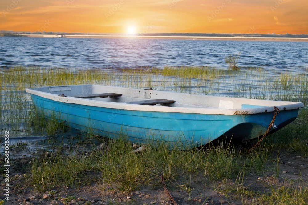 A blue small fishing boat moored on the bank of a dam in the reeds.Empty boat on the shore and a beautiful orange sunset. A blue small fishing boat moored on the bank of a dam in the reeds.Empty boat on the shore and a beautiful orange sunset.