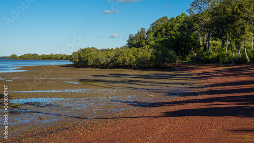Shadows on the red pebbly beach at low tide, with trees, mangroves and blue sea. Redland Bay, Queensland, Australia. 