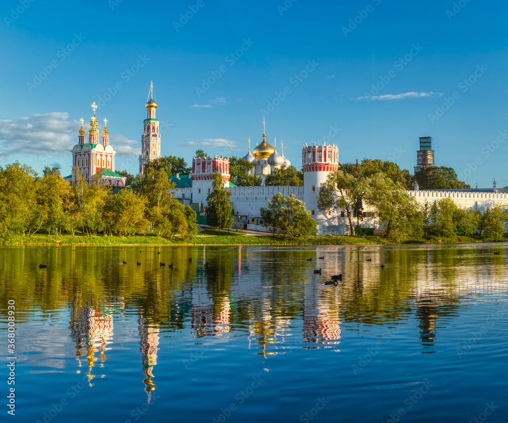 Obraz premium The famous Moscow landmark with Golden domes and a reflection of the landscape on the water of the Park pond against the background of a picturesque blue sky with clouds.