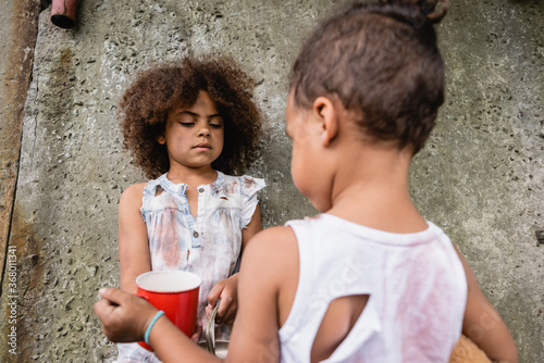 Selective focus of destitute afican american kid holding metal plate near brother and concrete wall on urban street