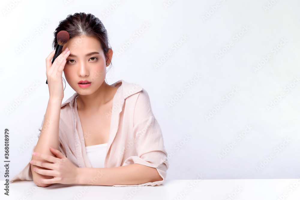 Portrait of beautiful charming young Asian woman brushing facial with cosmetic and sitting with white table, beauty of female face with skincare on white background, beauty cosmetic make up concept Portrait of beautiful charming young Asian woman brushing facial with cosmetic and sitting with white table, beauty of female face with skincare on white background, beauty cosmetic make up concept