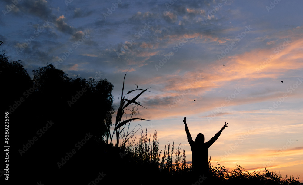A dark silhouette of a girl with raised hands against the background of the sunset.