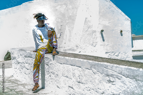 Italian immigrant man of sub-Saharan origin with sun hat and black and golden hair with an elegant look with the traditional colors of Ghana sitting on the wall of the belvedere of the white city