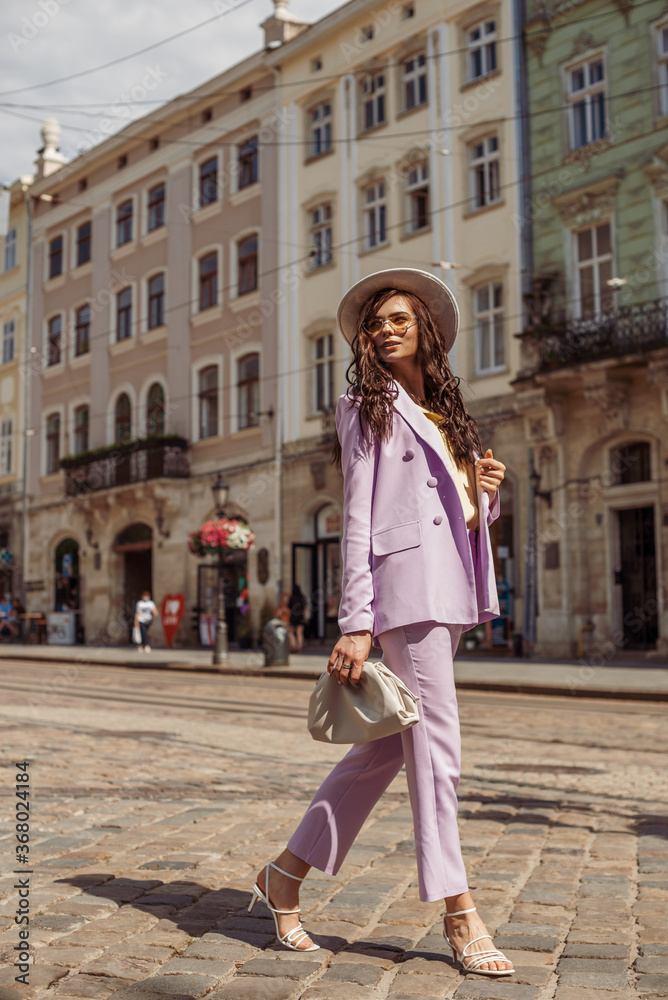 Outdoor full-length fashion portrait of elegant lady wearing lilac suit: double breasted  blazer, pants, strap sandals, holding trendy white leather pouch handbag, walking in street of European city