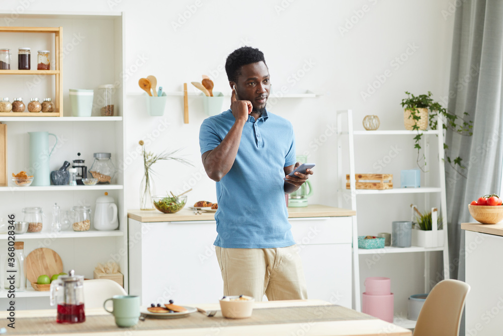 African young man using headphones to talk on mobile phone while standing in the kitchen at home African young man using headphones to talk on mobile phone while standing in the kitchen at home