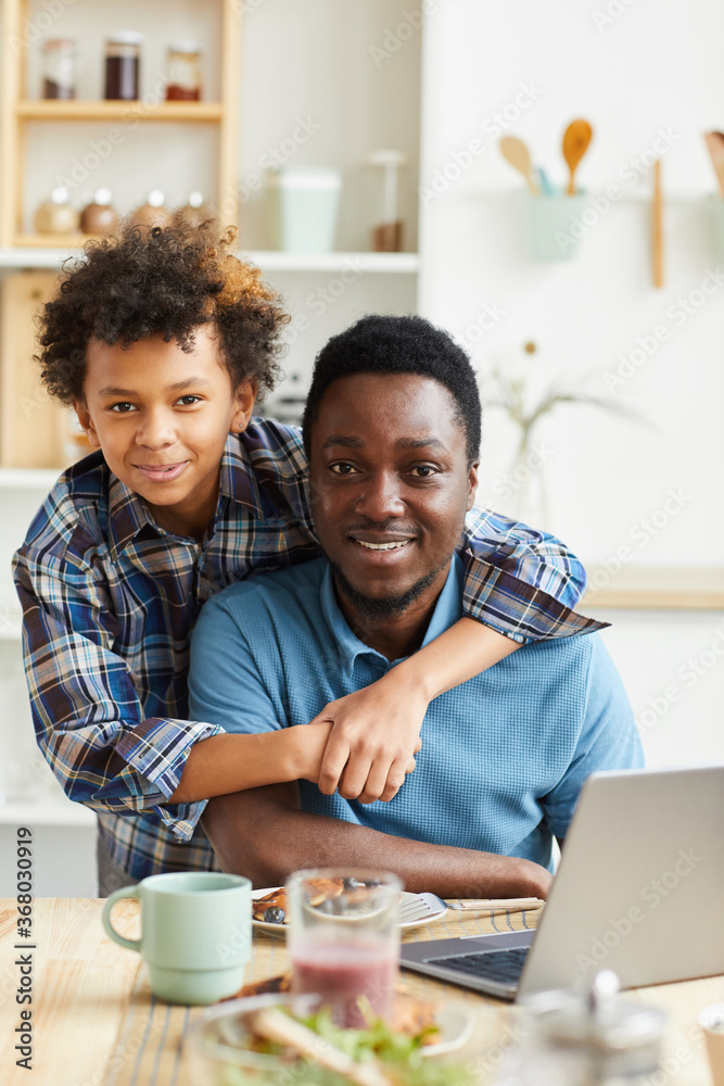 Portrait of happy father and son smiling at camera while sitting at the table with laptop in the kitchen Portrait of happy father and son smiling at camera while sitting at the table with laptop in the kitchen