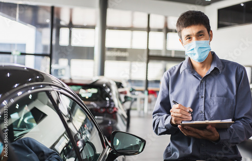 Asian saleman wear surgical mask working in inspector checking writing on clipboard in garage of dealership Wait mechanic In showroom car and insurance new normol covid-19 in showroom dealership photo