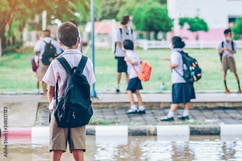 Male elementary school student wear face mask to prevent the Coronavirus(Covid-19) wait for her parents to pick her up to return home after school and the rain just stop in front of the school gate