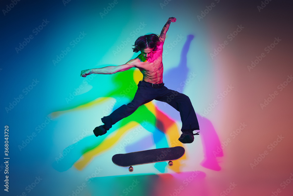 Skateboarder doing a trick isolated on studio background in colorful neon light. Young man shirtless riding and skateboarding in motion. Concept of leisure activity, sport, extreme, hobby and motion.