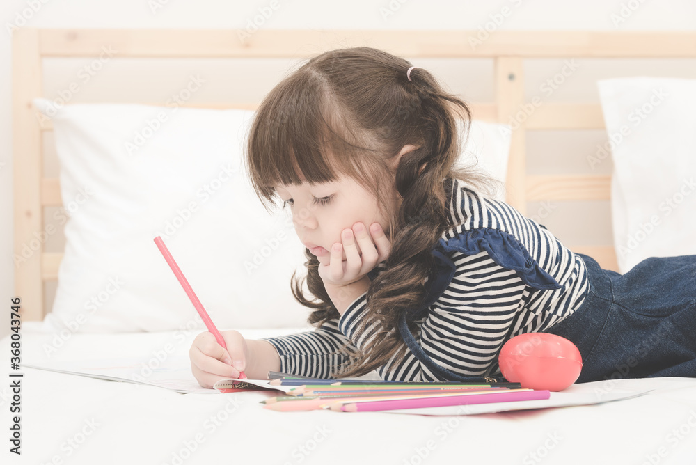 Cute asian little girl doing homework,writing with colourful pencils on bed at home. Elegant design for kid playing, preschool learning and creative art education concept. Cute asian little girl doing homework,writing with colourful pencils on bed at home. Elegant design for kid playing, preschool learning and creative art education concept.