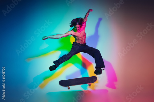 Skateboarder doing a trick isolated on studio background in colorful neon light. Young man shirtless riding and skateboarding in motion. Concept of leisure activity  sport  extreme  hobby and motion.