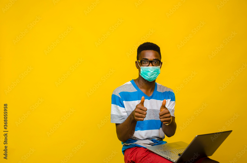 young handsome african man wearing face mask isolated over yellow background preventing himself from the outbreak in the society did thumbs up while his laptop is on his leg