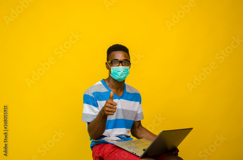 young handsome african man wearing face mask isolated over yellow background preventing himself from the outbreak in the society did thumbs up while his laptop is on his leg