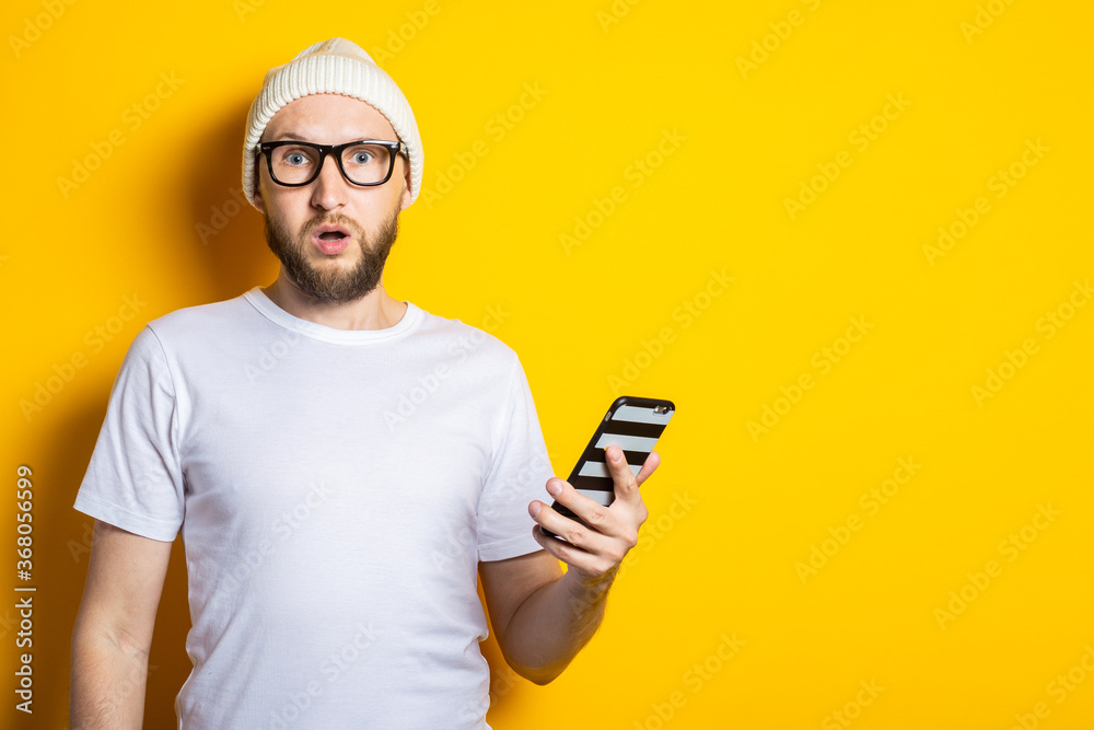 Surprised shocked young man with a beard holding a phone on a yellow background Surprised shocked young man with a beard holding a phone on a yellow background