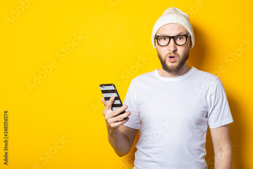 Surprised shocked young man with beard holding phone on yellow background.