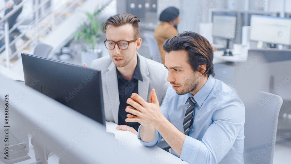 In Bright Modern Office: Handsome Office Worker Sitting and Working at His Desktop Computer, Discusses Problem Solution with His Male Project Manager. Diverse Team of Businesspeople Solving Problems In Bright Modern Office: Handsome Office Worker Sitting and Working at His Desktop Computer, Discusses Problem Solution with His Male Project Manager. Diverse Team of Businesspeople Solving Problems