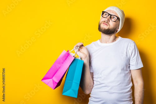 Young man with beard looking up and holding shopping bags on yellow background.