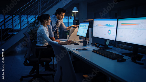 Valokuva Late at Night In Modern Office: Businessman and Businesswoman Work on Desktop Computer, Having Discussion, Finding Problem Solution, Finishing Project