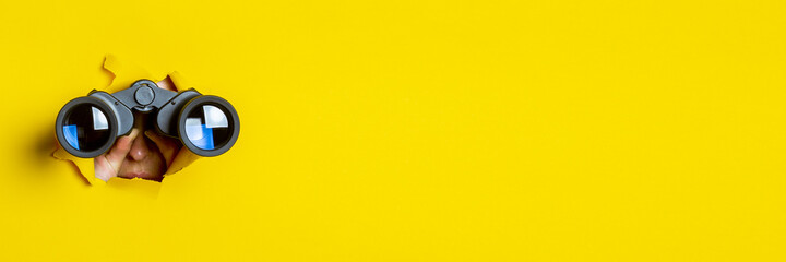 Female hand holds black binoculars on a yellow background. Journey, find and search concept. Banner.