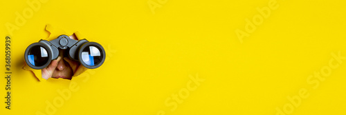 Female hand holds black binoculars on a yellow background. Journey, find and search concept. Banner.