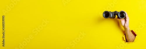 Female hand holds black binoculars on yellow background, travel, find and search concept. Banner photo