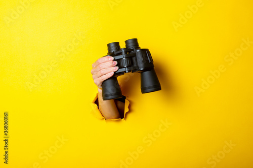 Female hand holds black binoculars on yellow background, travel, find and search concept