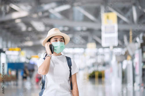 Young female wearing face mask and using mobile smartphone in airport terminal, protection Coronavirus disease (Covid-19) infection, Asian woman traveler with hat. New Normal and travel bubble concept
