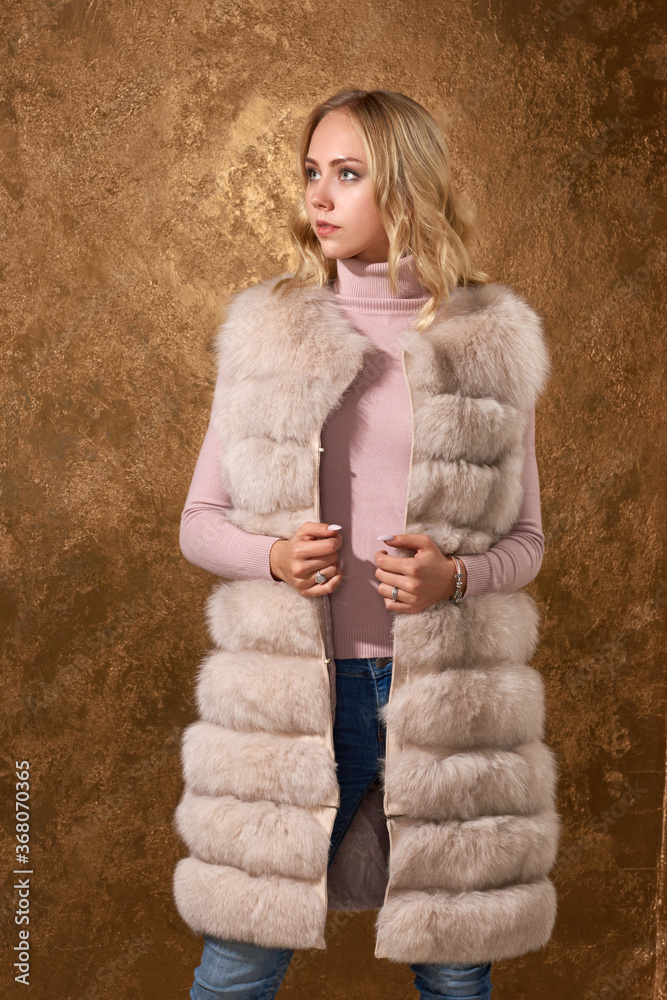 Luxury girl with long wavy blond hair in beige fur coat. Glamour woman posing against golden wall Luxury girl with long wavy blond hair in beige fur coat. Glamour woman posing against golden wall