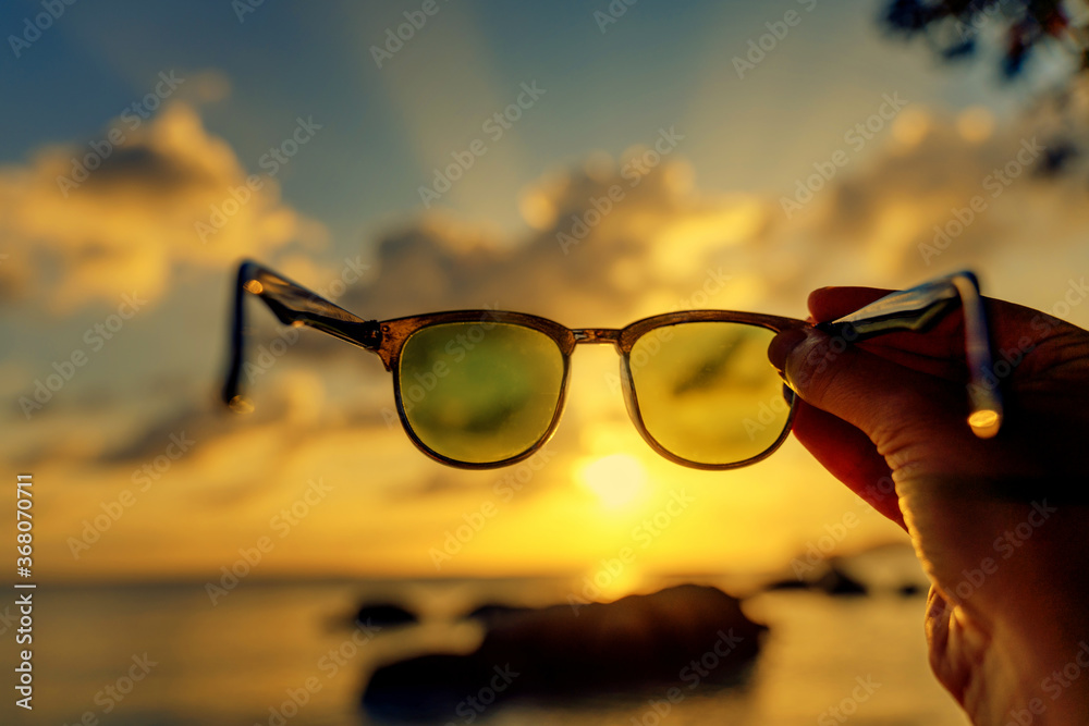 View of the sunset on the tropical sea through sunglasses in hand, abstract conceptual background of vacation and travel - obrazy, fototapety, plakaty 