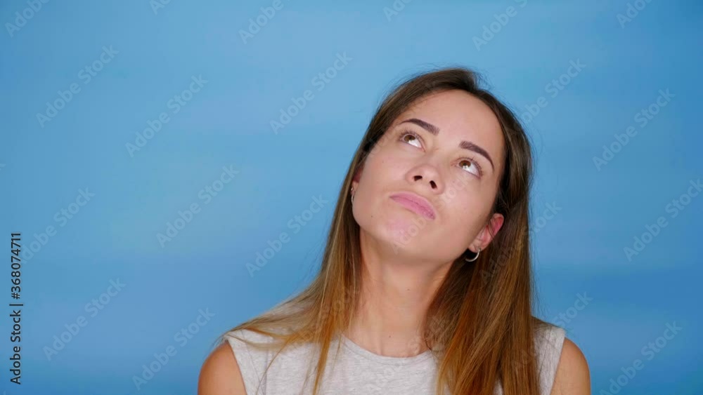 Beautiful woman in gray t-shirt looks up and thinks on blue background with copy space. 4K slow motion footage Beautiful woman in gray t-shirt looks up and thinks on blue background with copy space. 4K slow motion footage