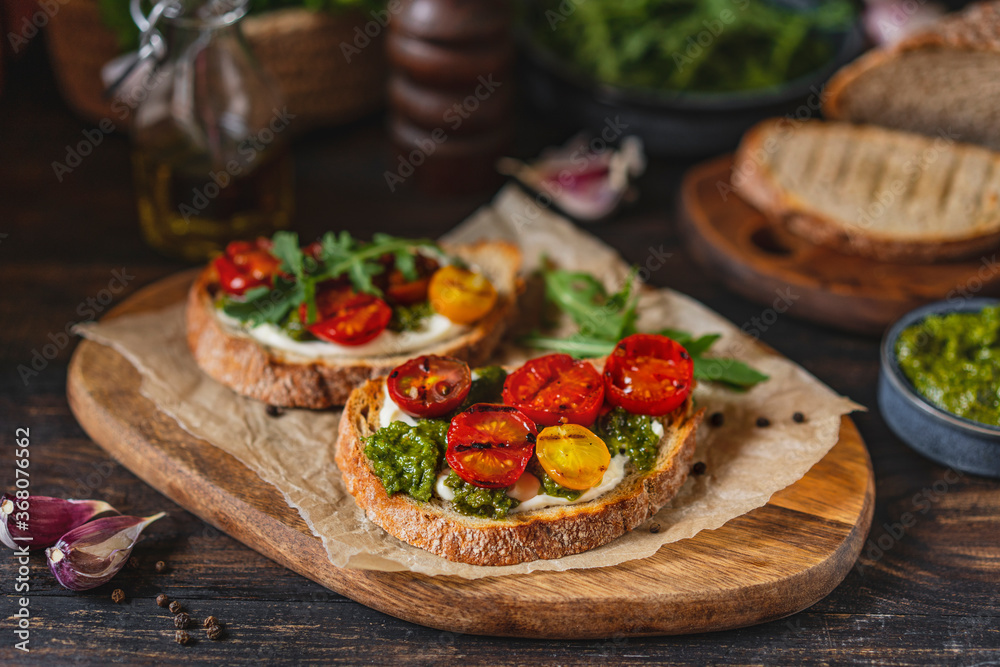 Bruschetta with cream cheese, pesto sauce and grilled cherry tomatoes, herbs on chopping board, rustic wooden table at home kitchen. Italian food, antipasto snack. Selective focus, copy space. Bruschetta with cream cheese, pesto sauce and grilled cherry tomatoes, herbs on chopping board, rustic wooden table at home kitchen. Italian food, antipasto snack. Selective focus, copy space.
