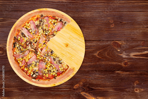Delicious pizza with chicken breast, corn, bacon and mushrooms, without a quarter on a round wood plate which is on wooden rustic table, top view and copy space