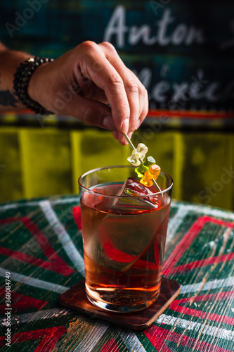 A transparent cocktail in a highball glass with a big ice cube. A hand putting gummy bears garnish on top of a drink