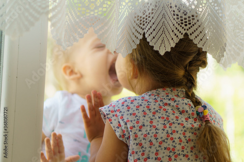 Little young smiling Toddler Child looks out window looking for his sister. Kids with small age difference play hide and seek. Brother and sister communicate and kiss through glass. Coronavirus, epide photo