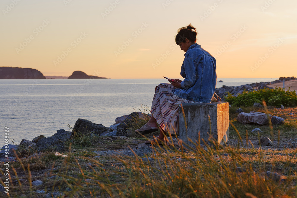 mujer lee un smartphone sentada en un banco junto a la playa al atardecer en asturias