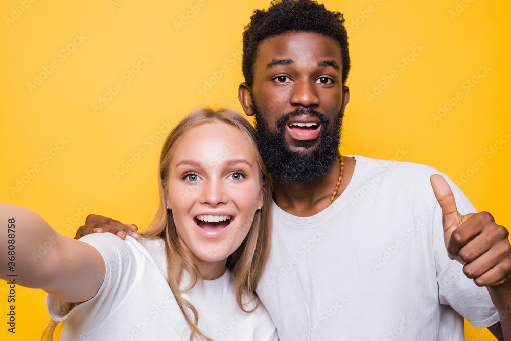 Obraz premium Selfie Fun. Cheerful interrational couple taking self-portrait together, looking at camera and smiling, posing over yellow background